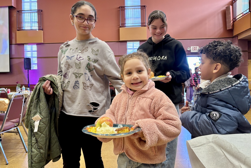 Several children and adults gather indoors, some holding plates of food.