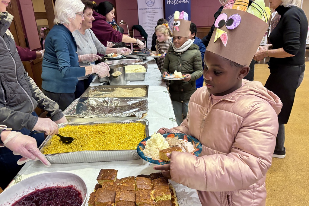 A child wearing a turkey hat holds a plate of food at a community gathering.