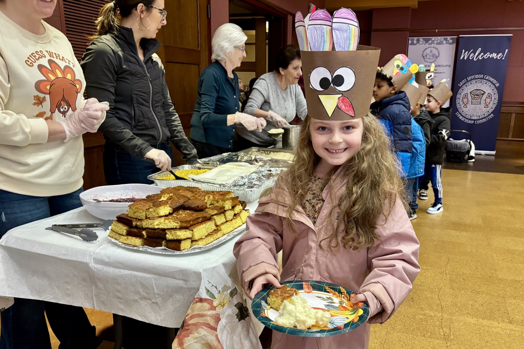 A young girl smiles, wearing a turkey hat and holding a plate of food at a Thanksgiving event.