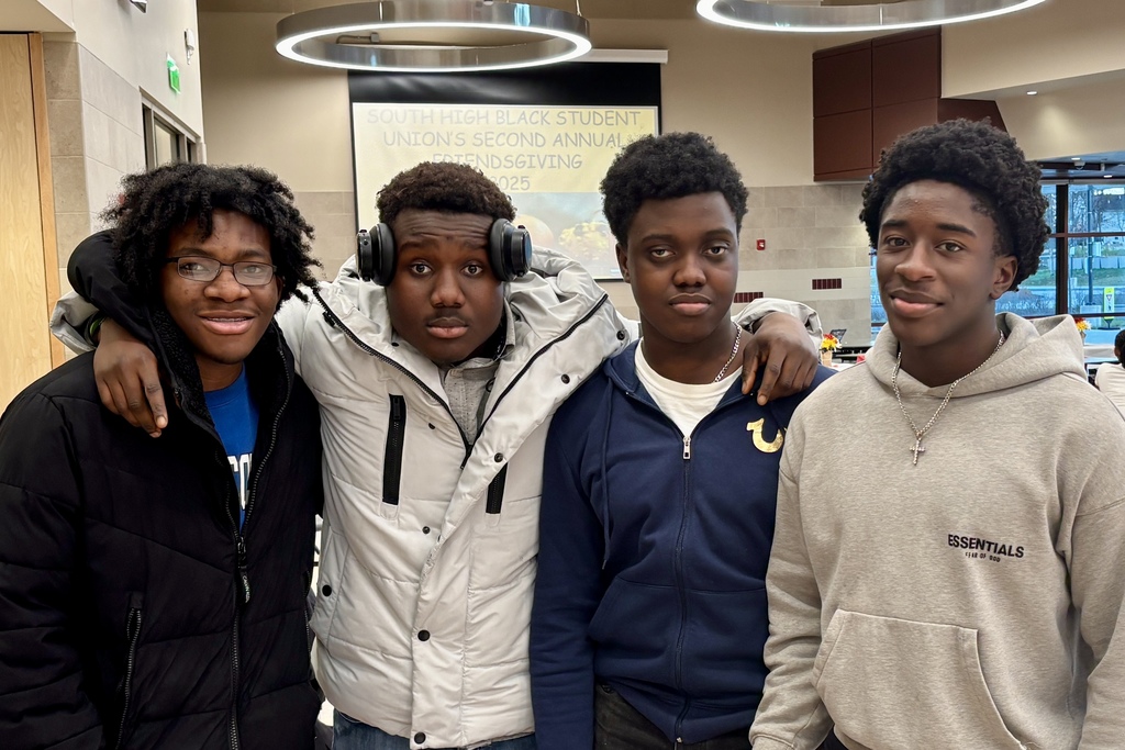 Four young men pose together, arms around each other, in a school setting.
