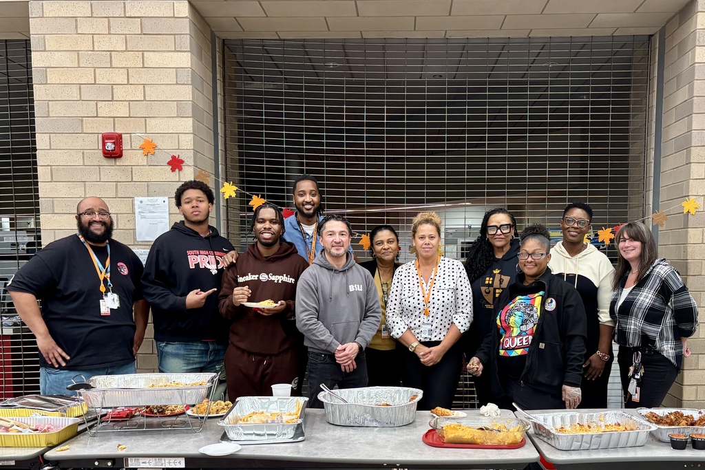 A diverse group of people stand behind a table laden with food, smiling at the camera.