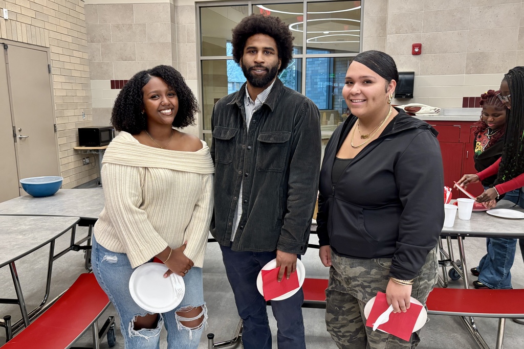 Three people smile, holding plates and red napkins in a cafeteria setting.