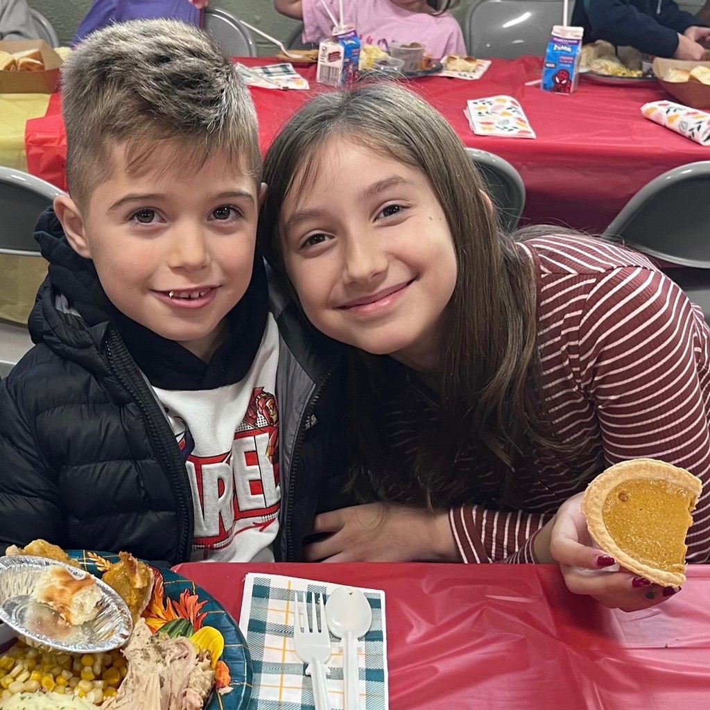 Two children smile at the camera while seated at a table with food