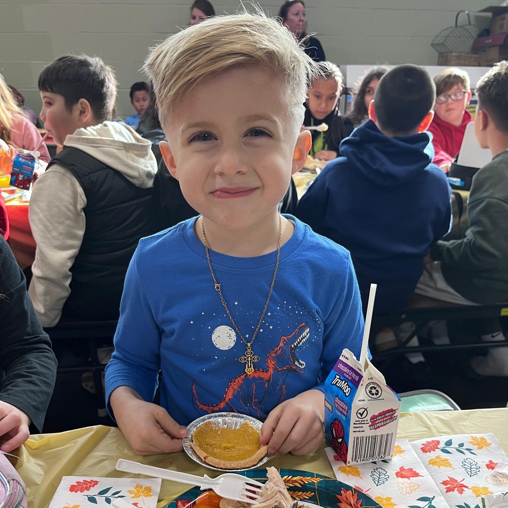 A young boy smiles at the camera while enjoying a meal at a table.
