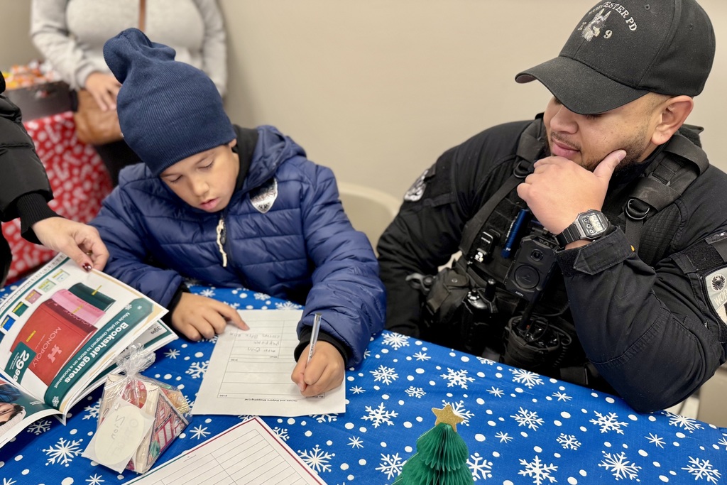 A child writes on a paper with a police officer, both at a table with a festive blue tablecloth.