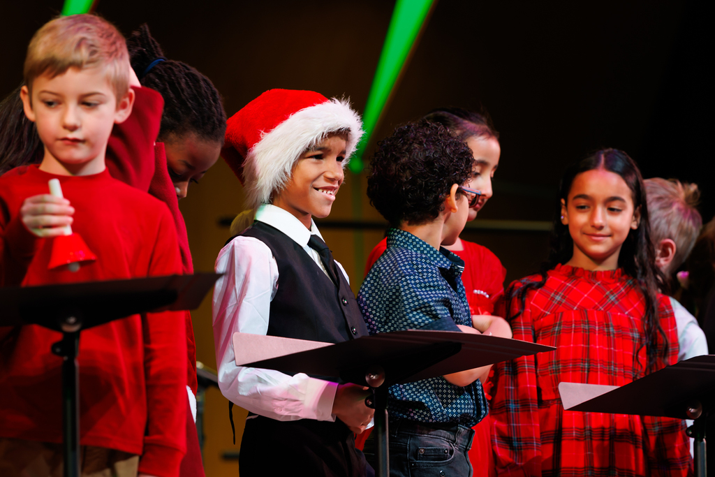 A young student wearing a santa hat smiles at his family in the crowd.