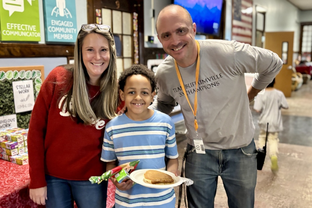 A smiling woman, a young boy holding a pancake, and a man in a grey shirt stand together indoors.