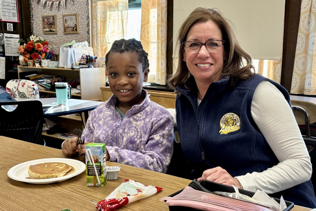A young girl with braided hair smiles brightly while sitting at a table with a woman wearing glasses.