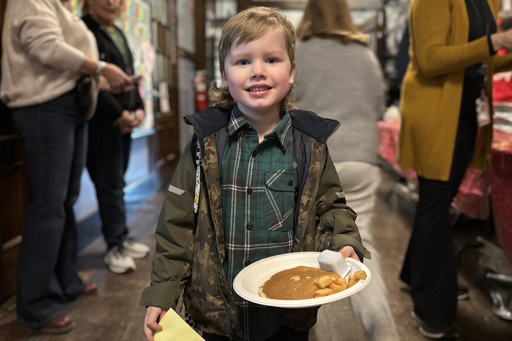 A young boy smiles while holding a plate of pancakes and fries at an indoor event.