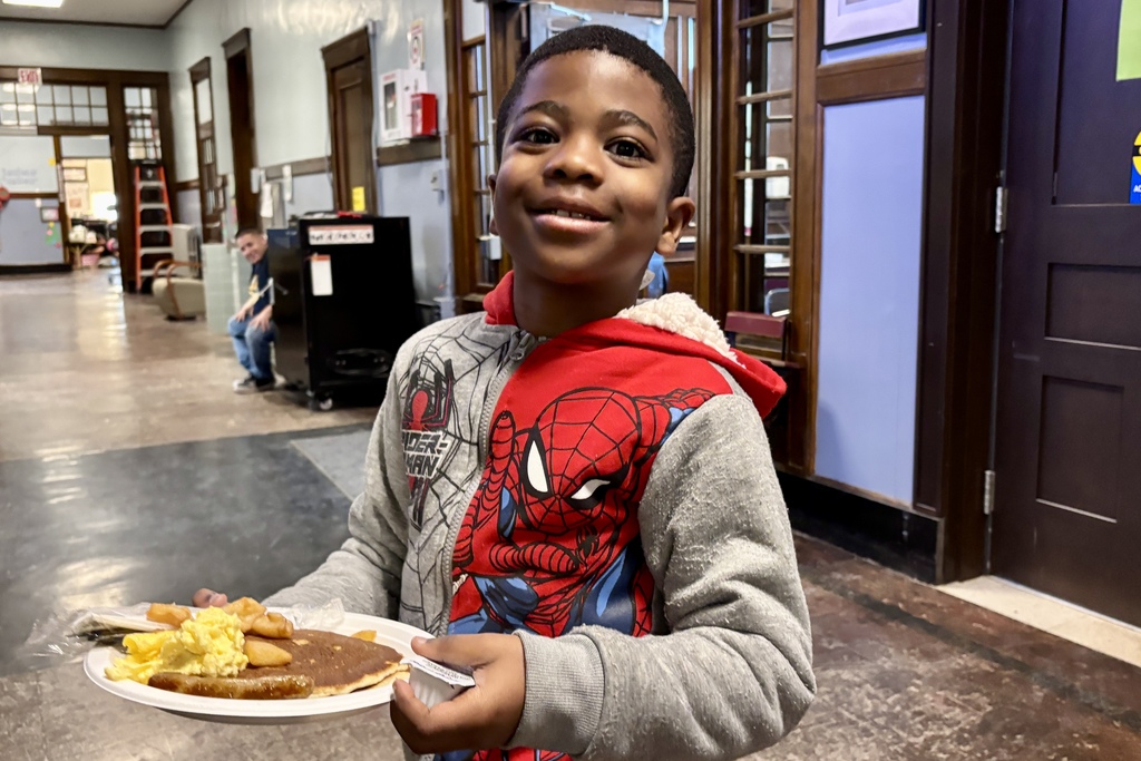 A young boy in a Spider-Man hoodie smiles while holding a plate of pancakes, scrambled eggs, and a sausage.