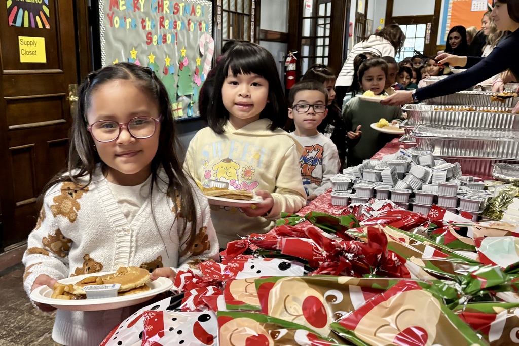 Children with plates of food at a holiday event, with festive decorations in the background.
