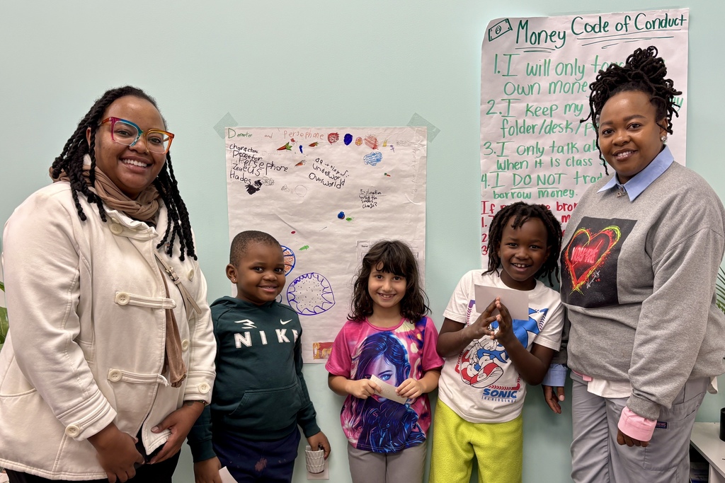 Two adults and three children stand in front of a wall with colorful drawings and a "Money Code of Conduct" poster.