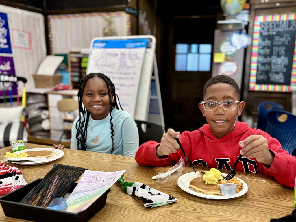 Two children smile while eating breakfast at a wooden table in a classroom.
