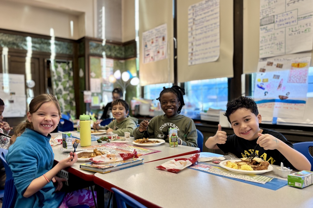 Four smiling children sit at a table in a classroom, enjoying a meal together.