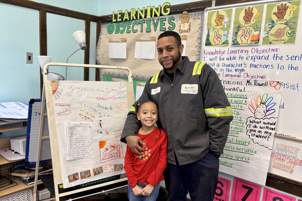 A man in a work jacket and a young girl stand smiling in a classroom with learning objectives displayed on the wall.