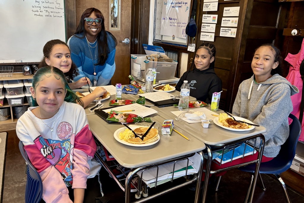 A teacher smiles as students eat breakfast at their desks in a classroom.