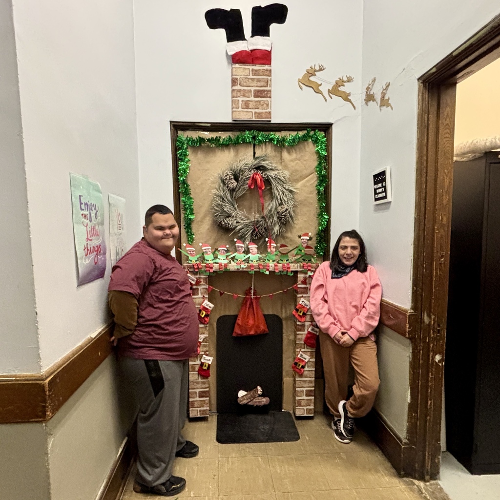Two smiling individuals stand in a hallway decorated with a festive Christmas fireplace, complete with stockings and a wreath.