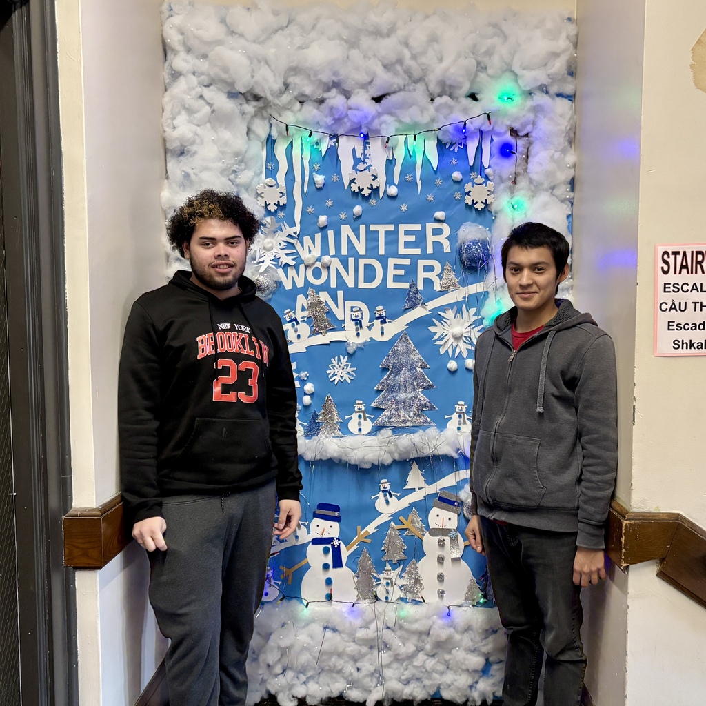 Two young men stand in front of a festive "Winter Wonder" door decoration featuring snowmen, trees, and snowflakes.