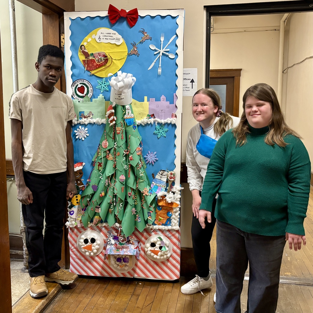 Three young people stand next to a festive, food-themed Christmas bulletin board display.