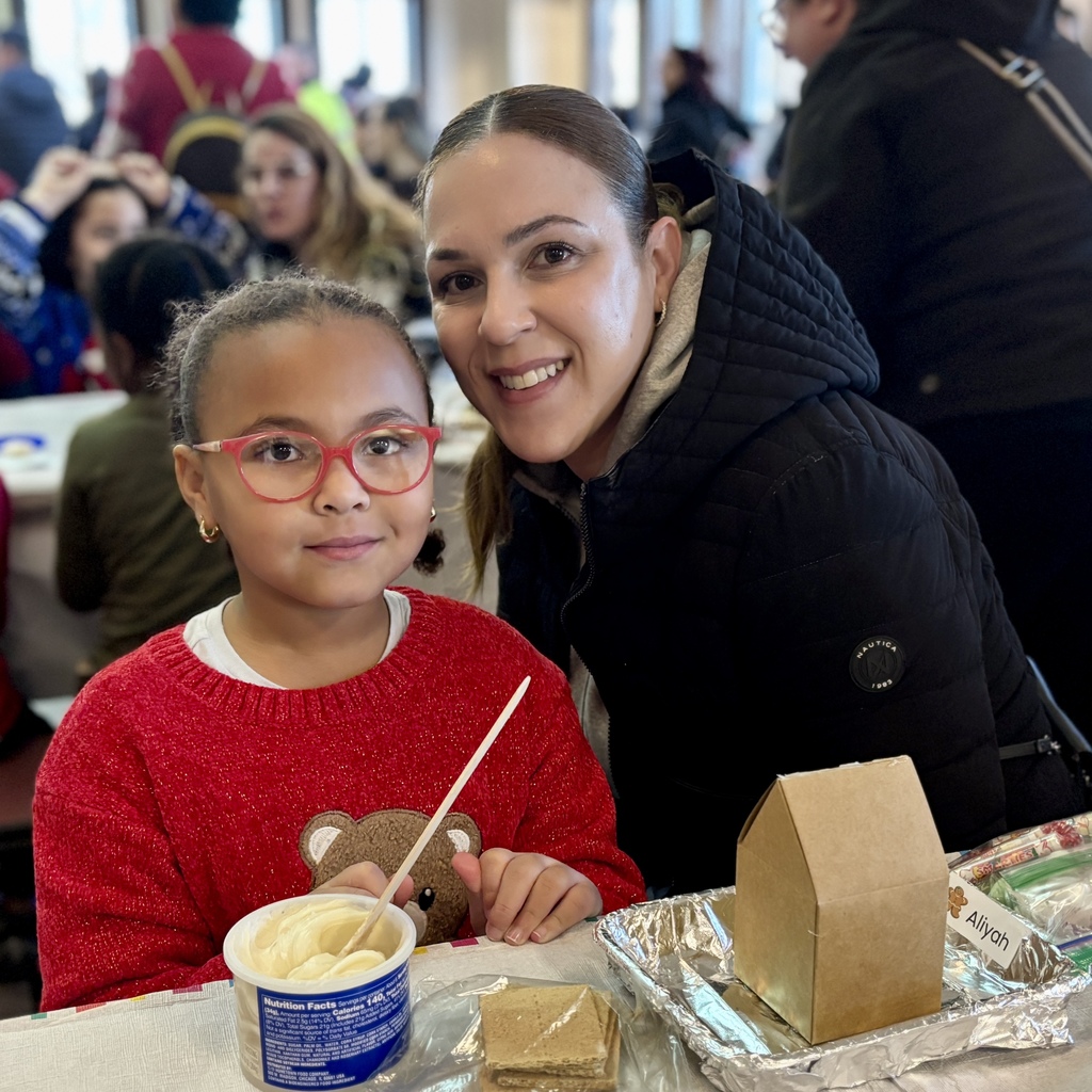A woman and a young girl smile at the camera while preparing to decorate a gingerbread house.