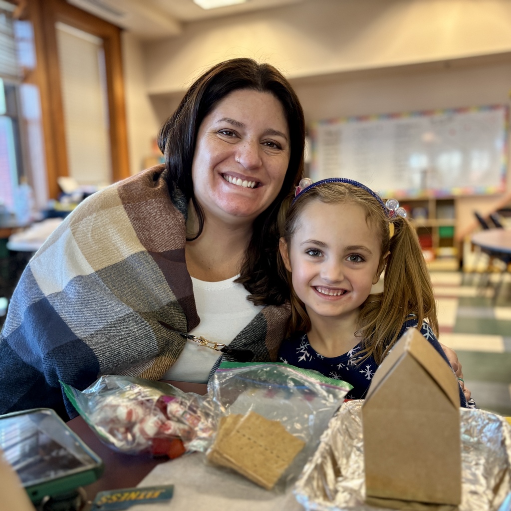 A woman and a young girl smile at the camera, with gingerbread house building supplies on the table.