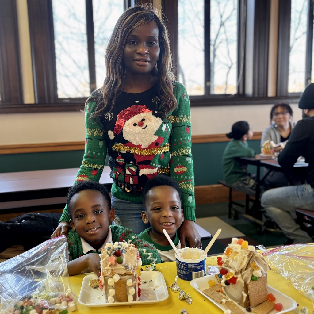 A woman in a festive Santa sweater smiles with two young boys, surrounded by decorated gingerbread houses.
