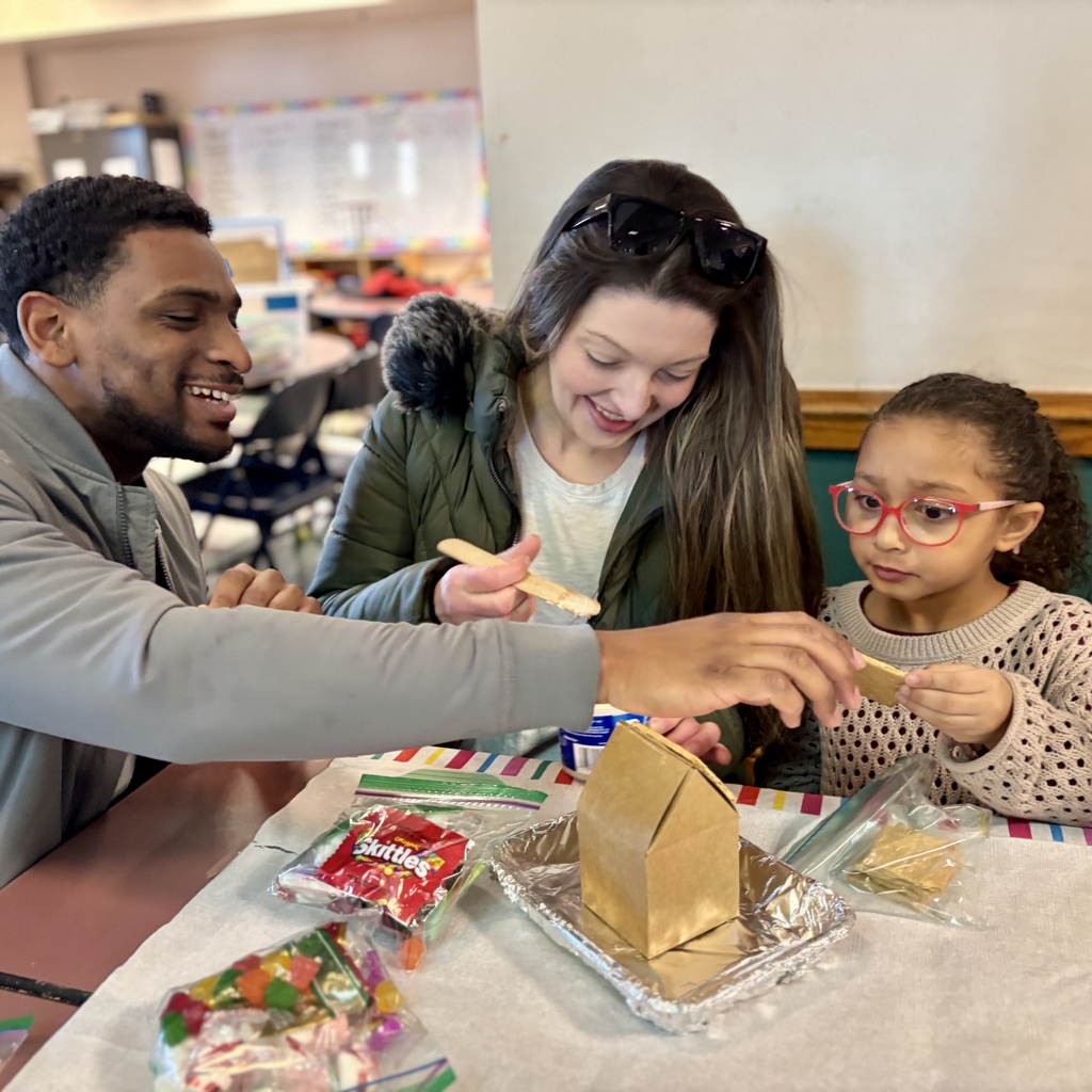 A family smiles while decorating a gingerbread house with candy and frosting.