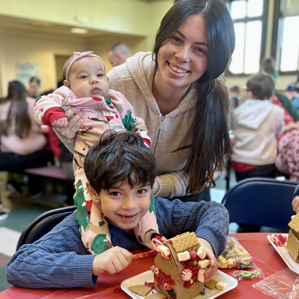 A smiling boy decorates a gingerbread house with candy while his mother holds a baby.