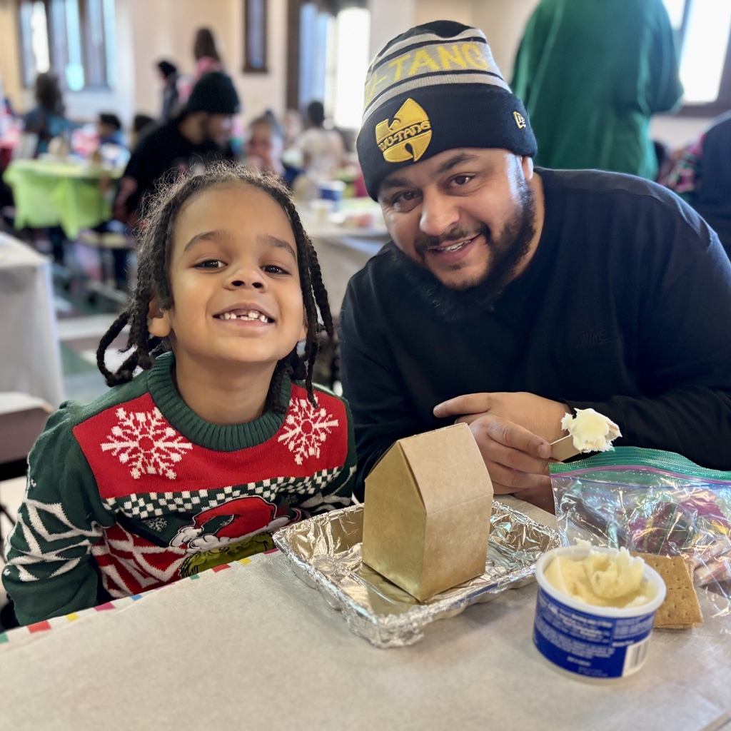 A man and a young child smile at the camera while working on a gingerbread house.