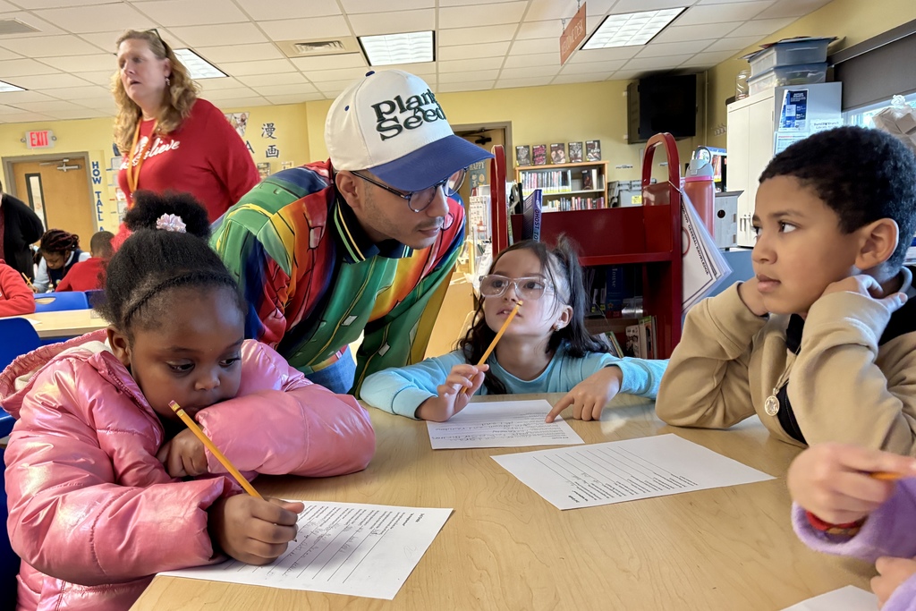 A teacher in a red shirt observes students working at desks in a classroom.