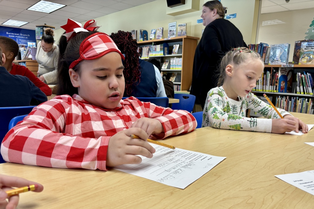 Children focus on writing assignments at wooden desks in a classroom with bookshelves.