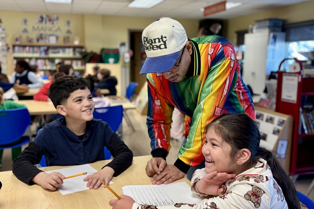 A teacher in a colorful jacket and a 'Plant Seed' hat interacts with two smiling students at their desks.