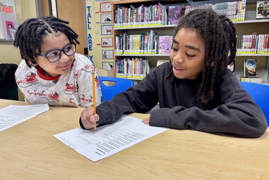 Two children are working on schoolwork at a wooden desk, with bookshelves in the background.