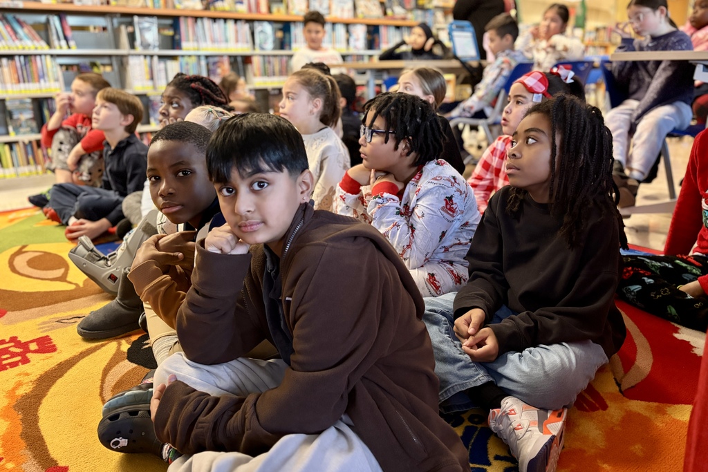 Children sit on a colorful rug in a library, looking towards the front.
