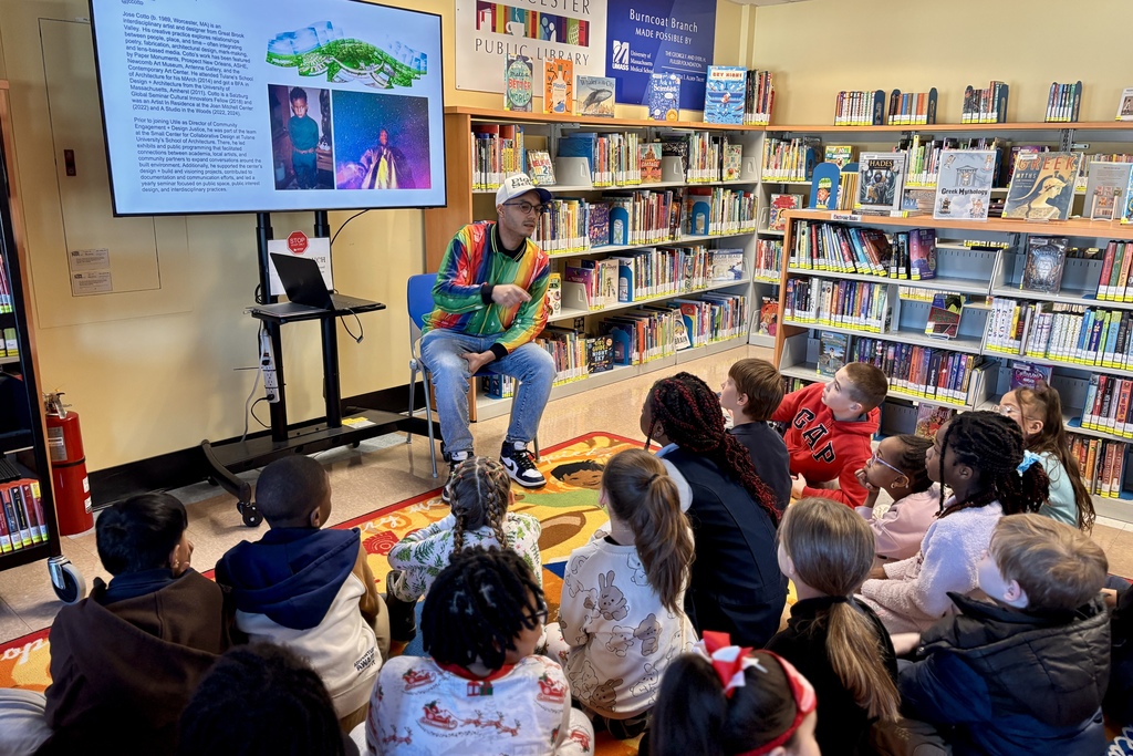 A presenter in a colorful jacket speaks to a group of children sitting on a rug in a library, with bookshelves in the background.