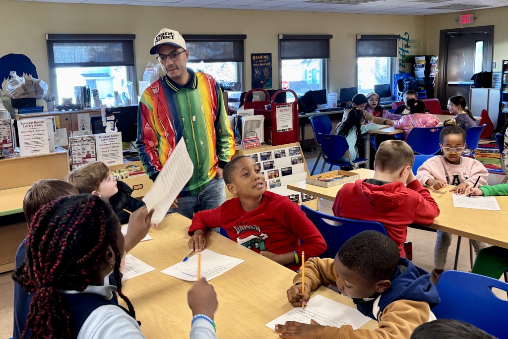 A teacher in a colorful jacket stands near a group of elementary school students working at desks in a classroom.