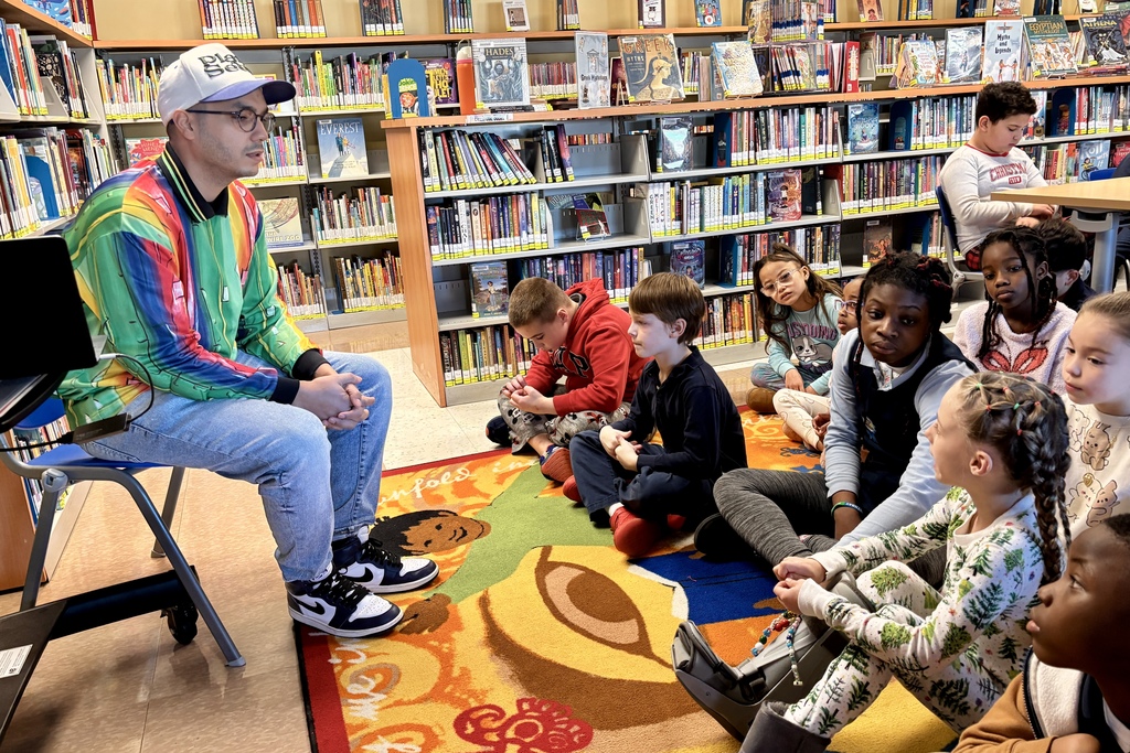 A man in a colorful jacket sits on a chair, facing a group of children seated on a rug in a library.