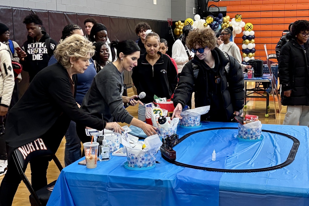 A group of students and adults gather around a table with a model train set and various items in decorative containers.