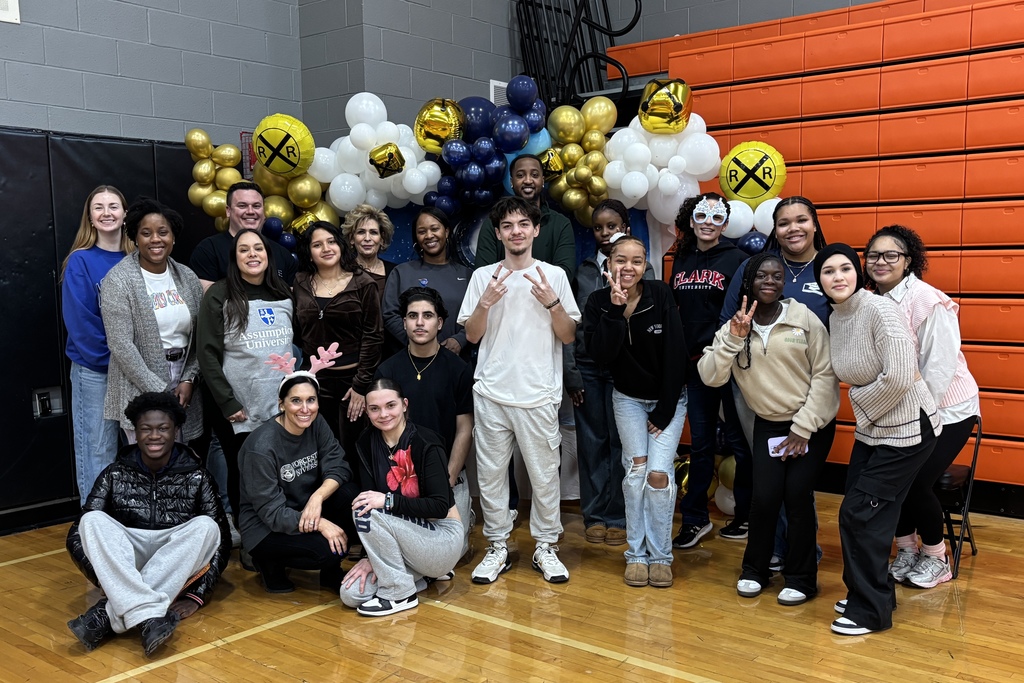 A diverse group of people, including students and adults, pose for a photo in a gymnasium with balloons.