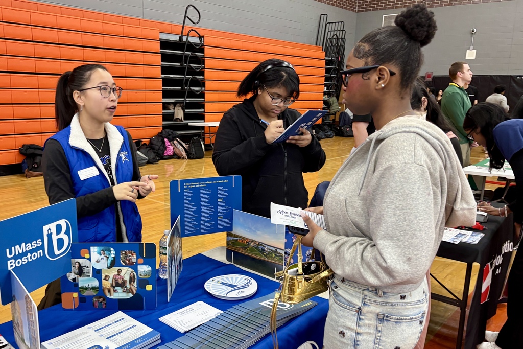 A UMass Boston representative in a blue vest speaks with students at a college fair booth.