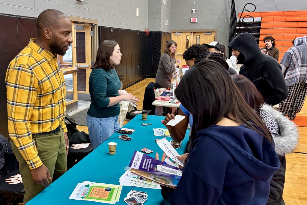 A diverse group of people gather around a teal table at an indoor event, with flyers and brochures spread out.