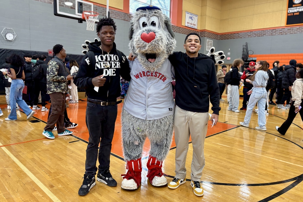 Two young men pose with a furry mascot wearing a 'Worcester' baseball jersey on a basketball court.