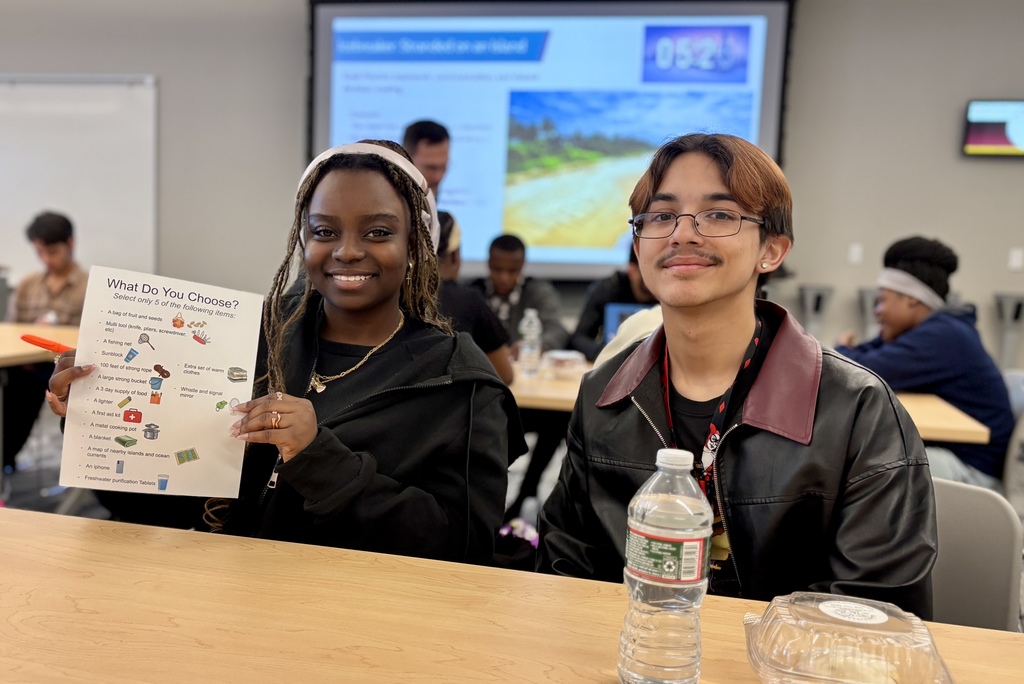 Two students, a young woman and a young man, smile at the camera in a classroom setting, holding a worksheet titled 'What Do You Choose?'.