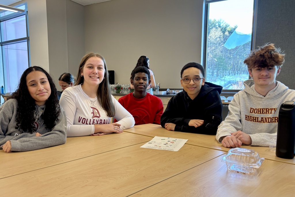 Five students sit at a wooden table in a classroom, with a large window showing a snowy outdoor scene.