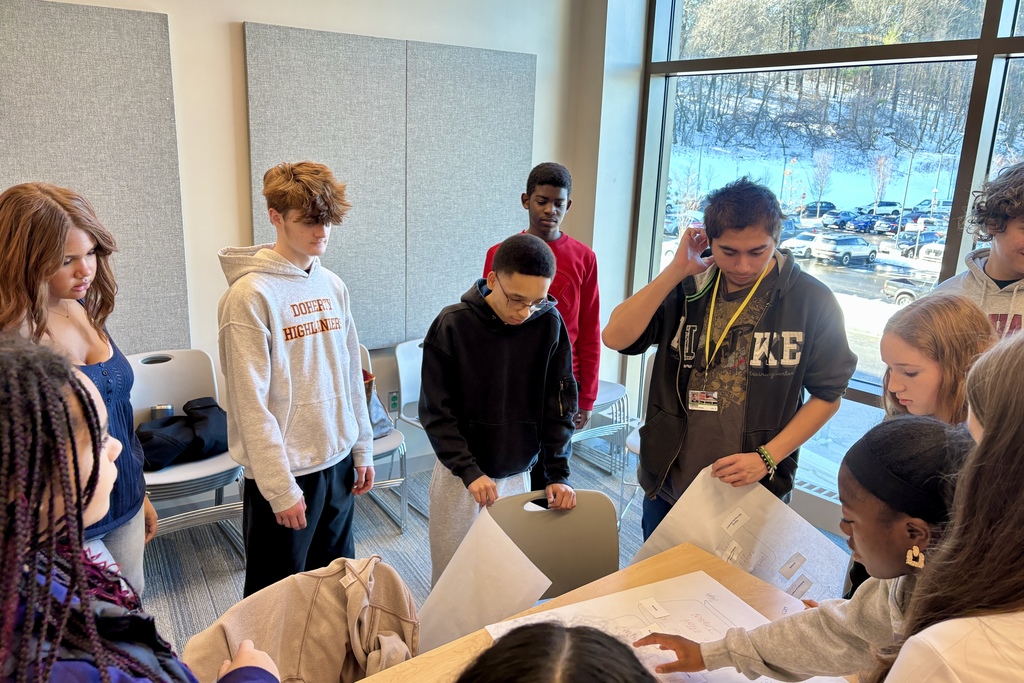 A diverse group of students collaborates around a table, reviewing papers and diagrams.