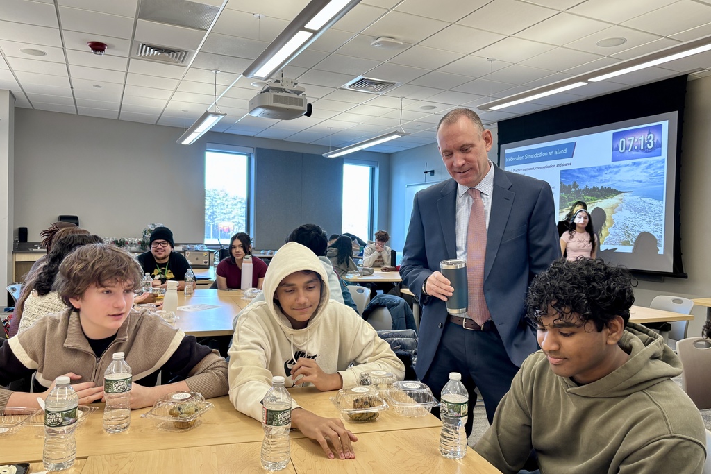 A man in a suit stands near a projector screen displaying an icebreaker activity titled 'Stranded on an Island' to a group of students.