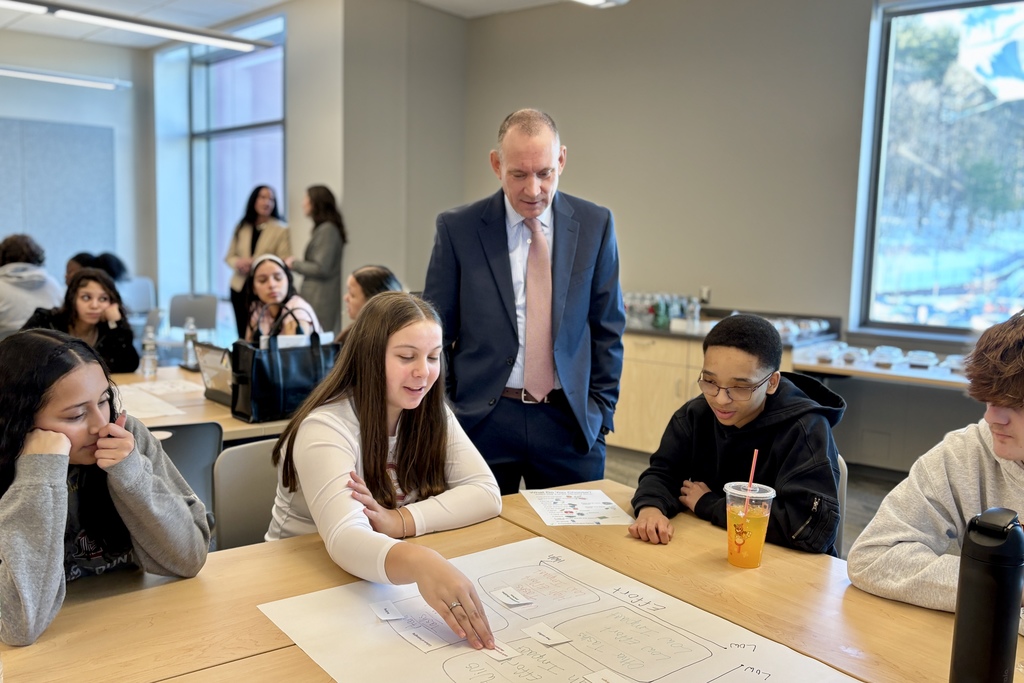 A group of students and an adult in a suit are gathered around a table, looking at a diagram on paper.