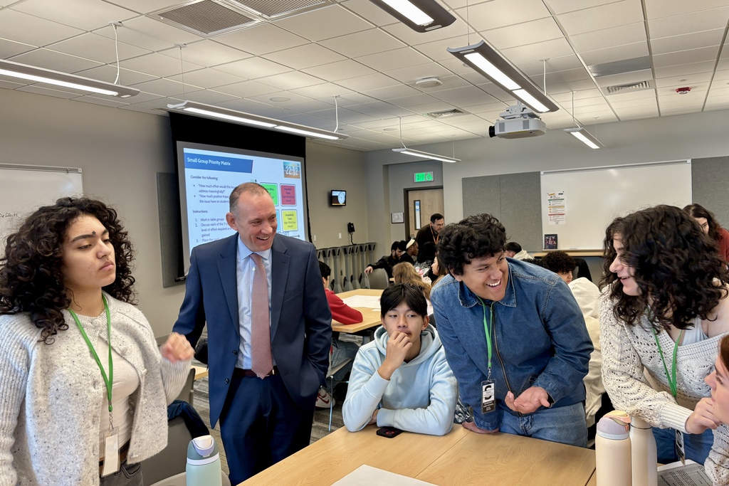 A teacher in a suit smiles while interacting with students in a classroom.