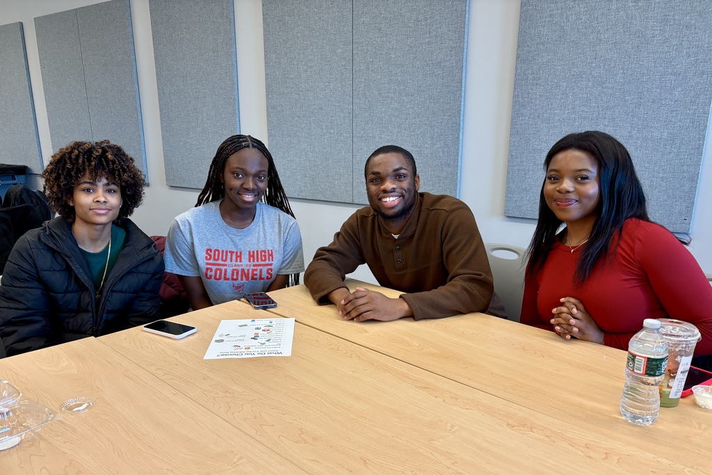 Four young people smile while sitting at a wooden table in a room with acoustic paneling.
