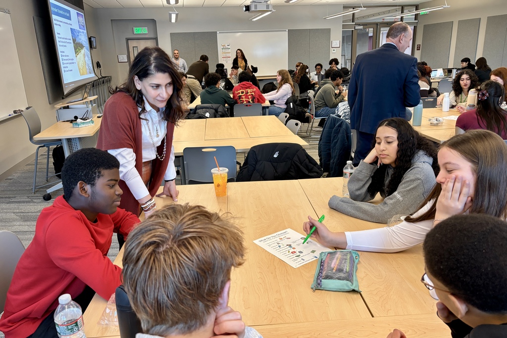 Students and adults gather around tables in a classroom, engaged in a group activity.